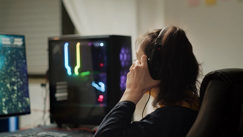 Achteraanzicht van een vrouwelijke gamer die een koptelefoon draagt en naar haar computer monitors kijkt met een kleurrijke RGB-verlichte pc-behuizing in de buurt.